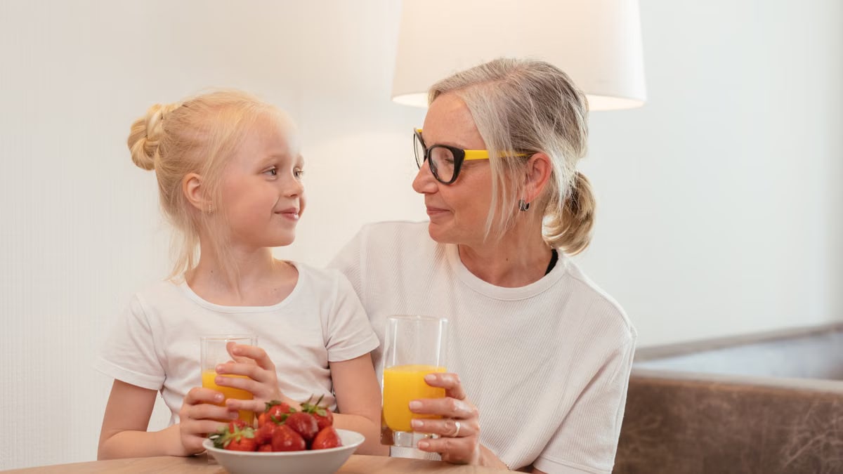 Elderly woman and young girl enjoying orange juice and strawberries at home, sharing a joyful moment together.